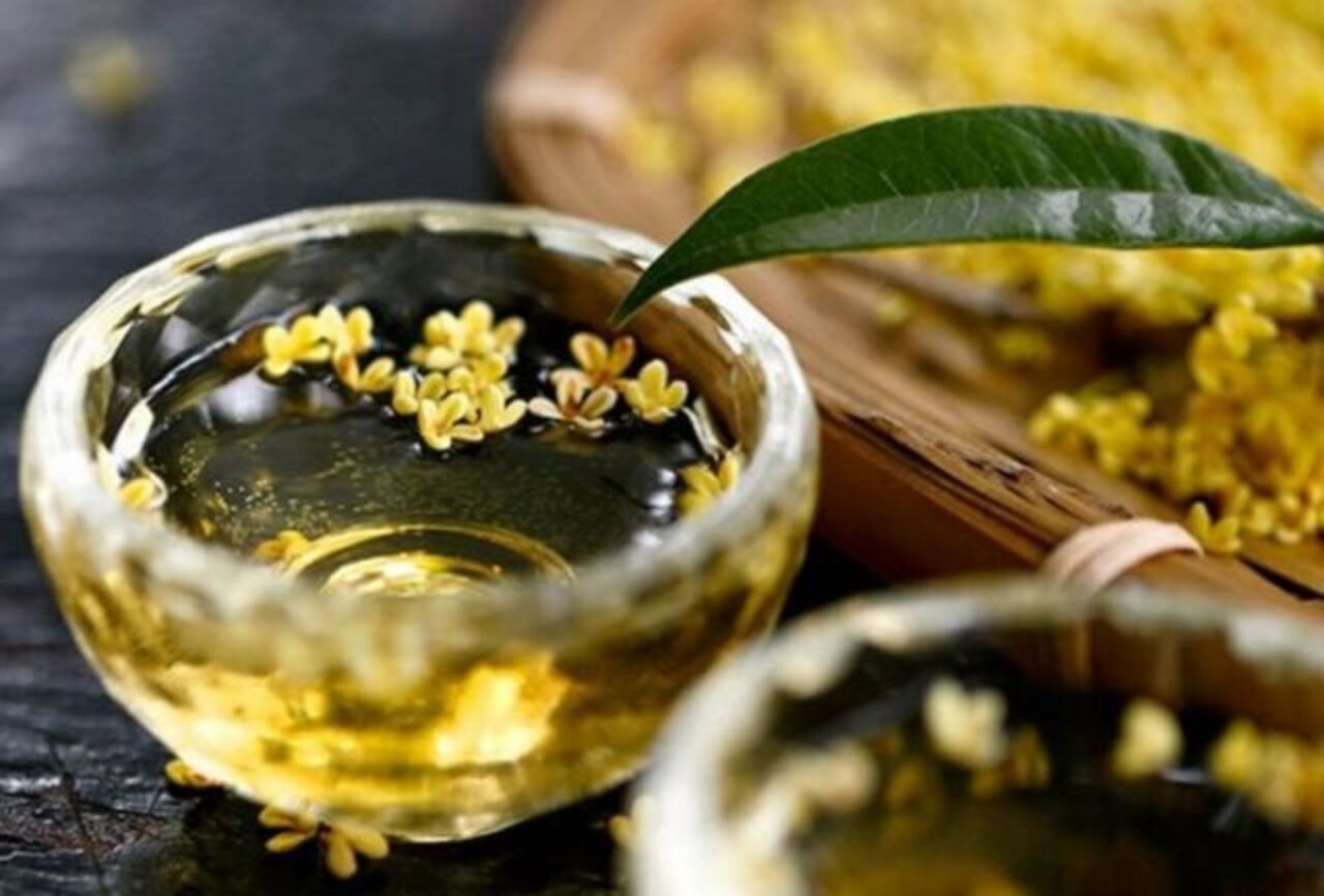 Un tazón de vidrio transparente contiene un líquido amarillo con pequeñas flores amarillas, una cuchara de madera y una hoja verde.