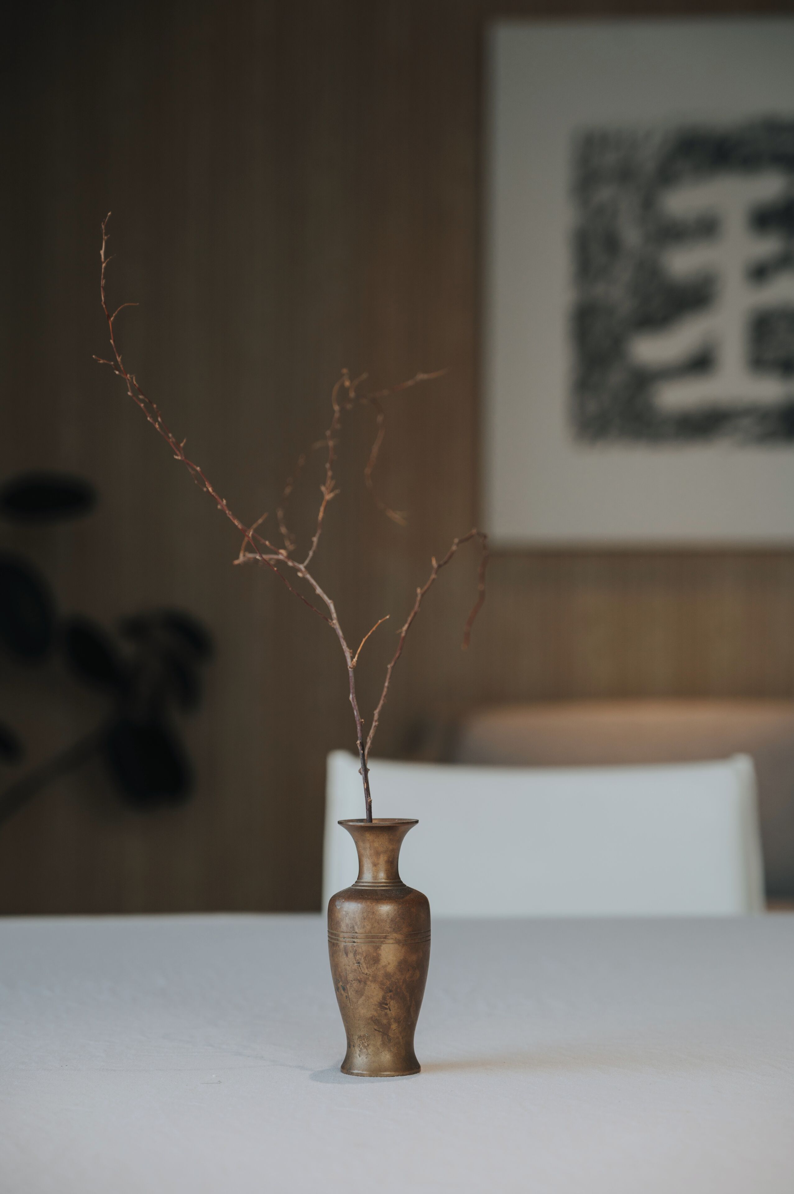A vase with branches on a table in a room with a bed and a framed picture on the wall.