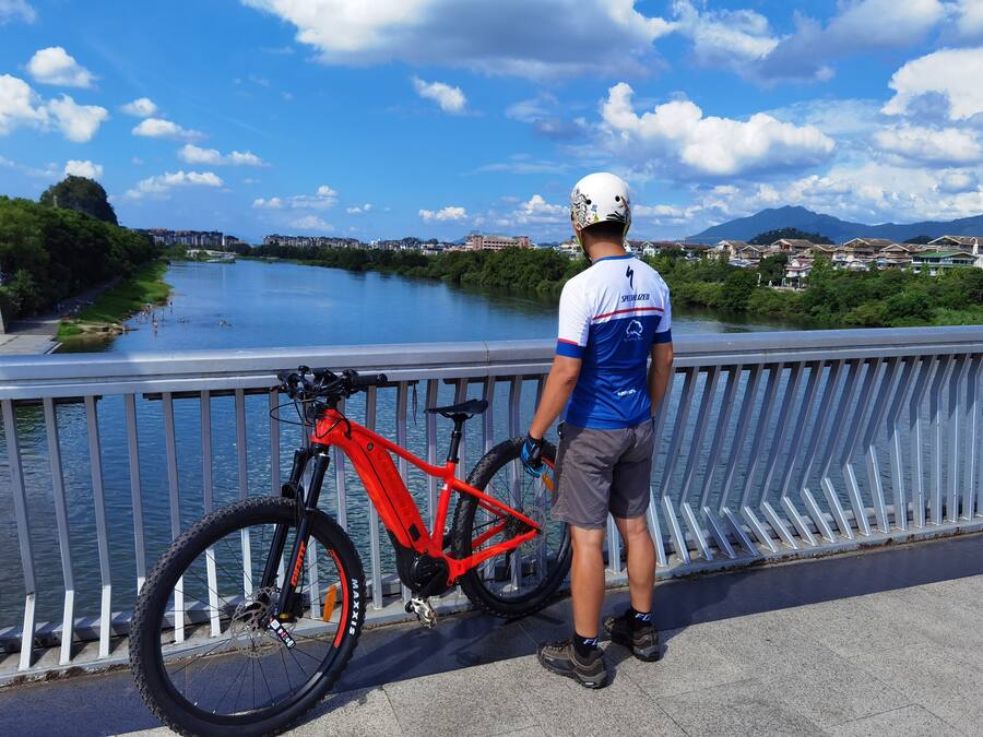 A person standing on a bridge with a red bicycle, overlooking a river and cityscape.