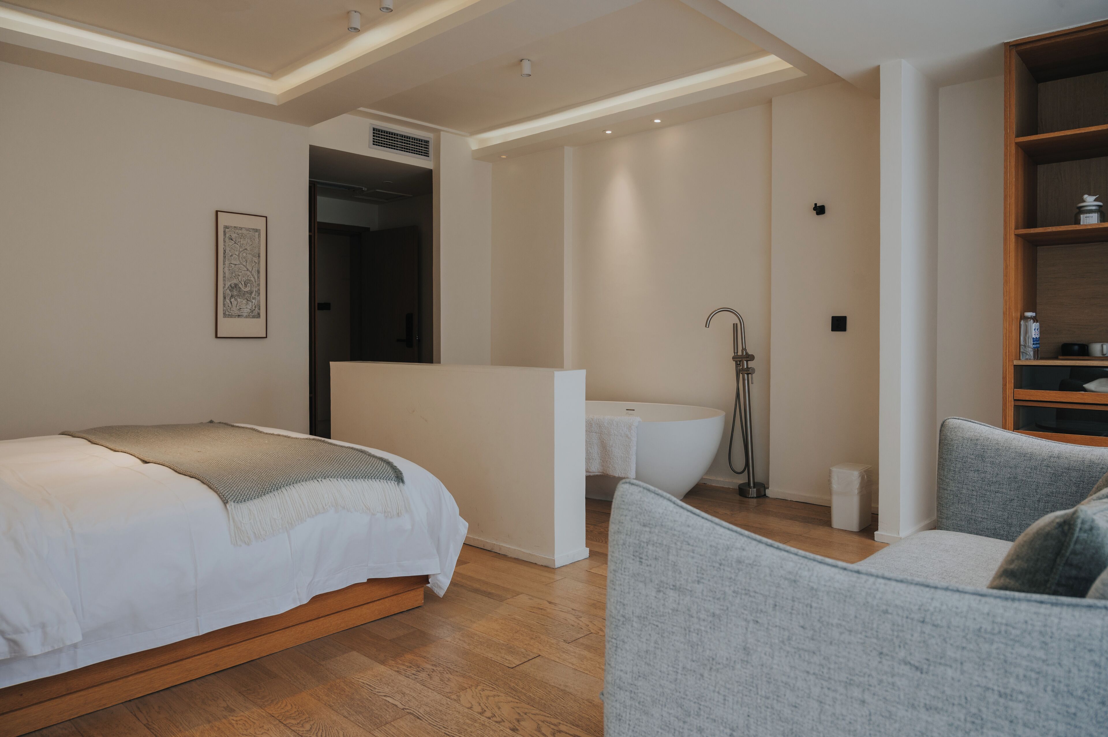 A modern bedroom with a bed, a gray armchair, a white bathtub, and a wooden shelf.