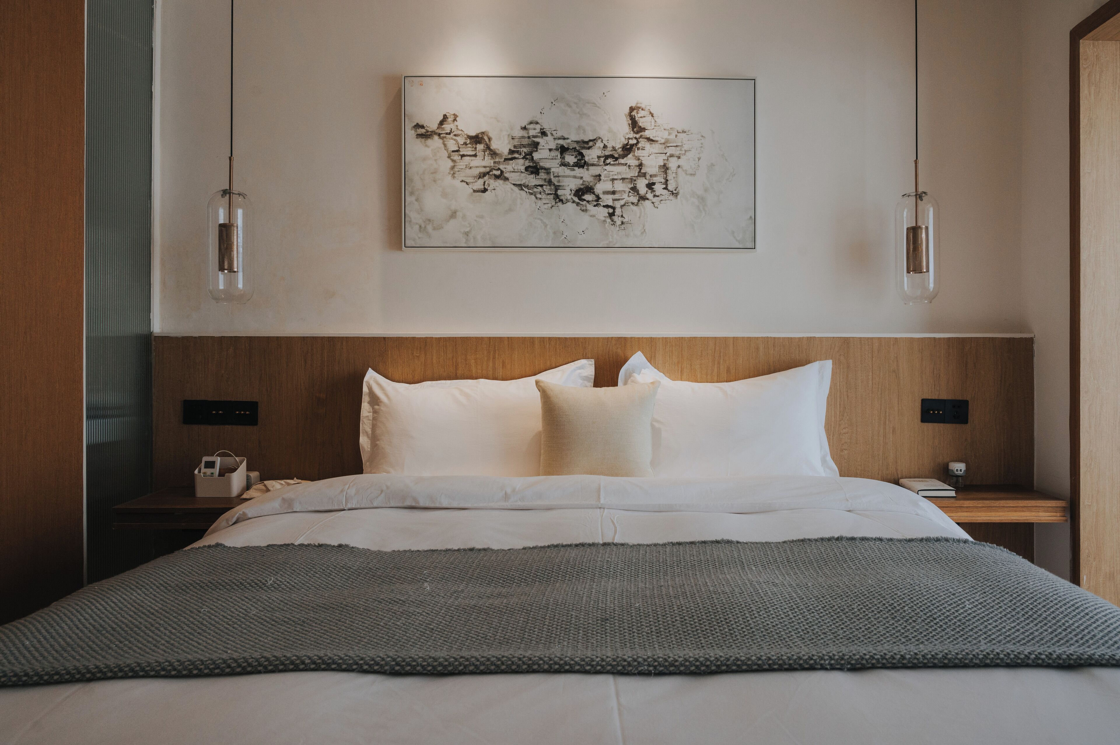 Una habitación de hotel moderna con cabecera de madera, una cama grande con sábanas blancas, una manta gris y una obra de arte abstracta colgada en la pared.