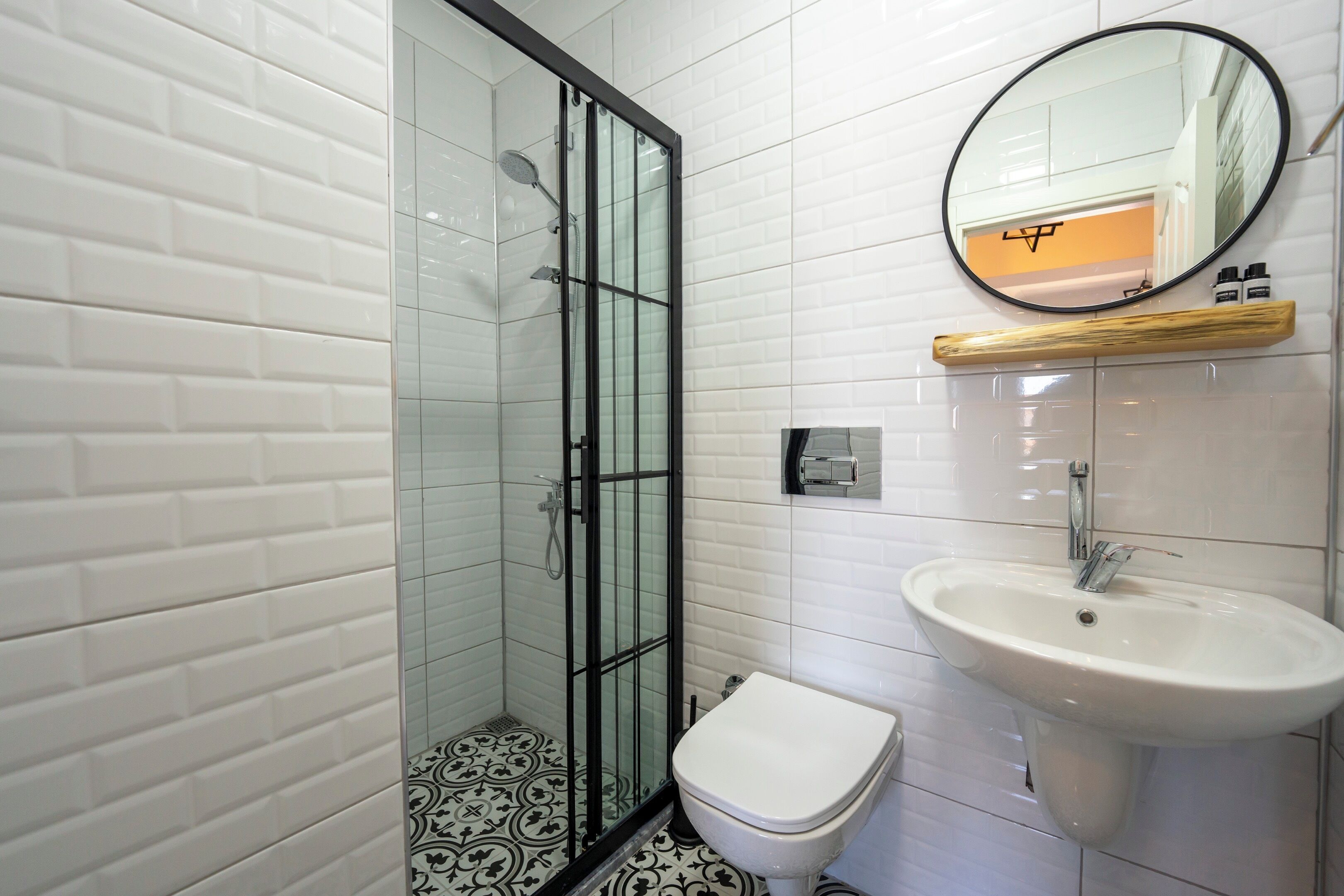 Un baño con paredes de azulejos blancos, ducha con mampara de vidrio, inodoro blanco y lavabo con espejo.