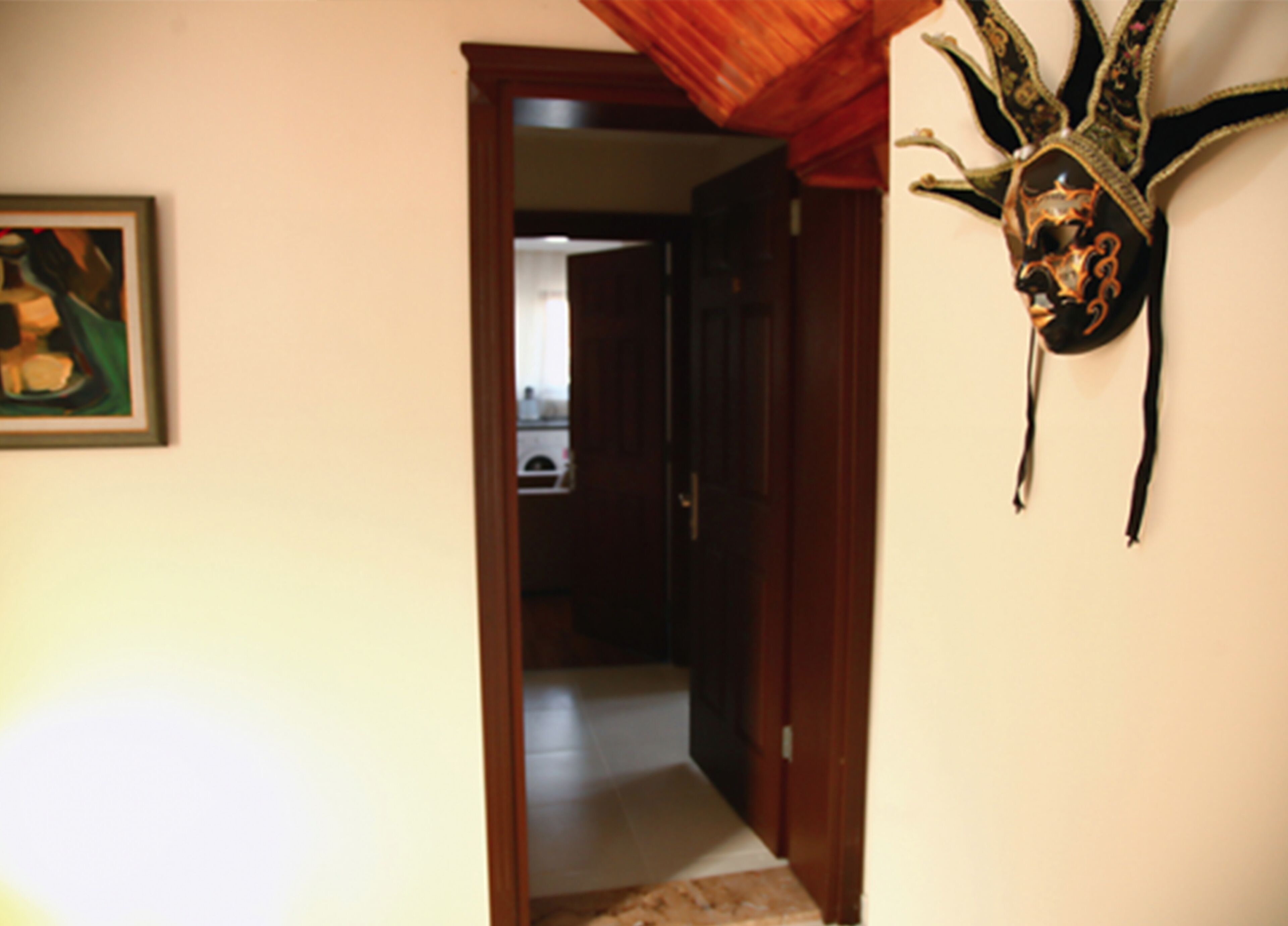 A hallway with a wooden door leading to a kitchen, a framed artwork on the wall, and a decorative mask hanging on the right.