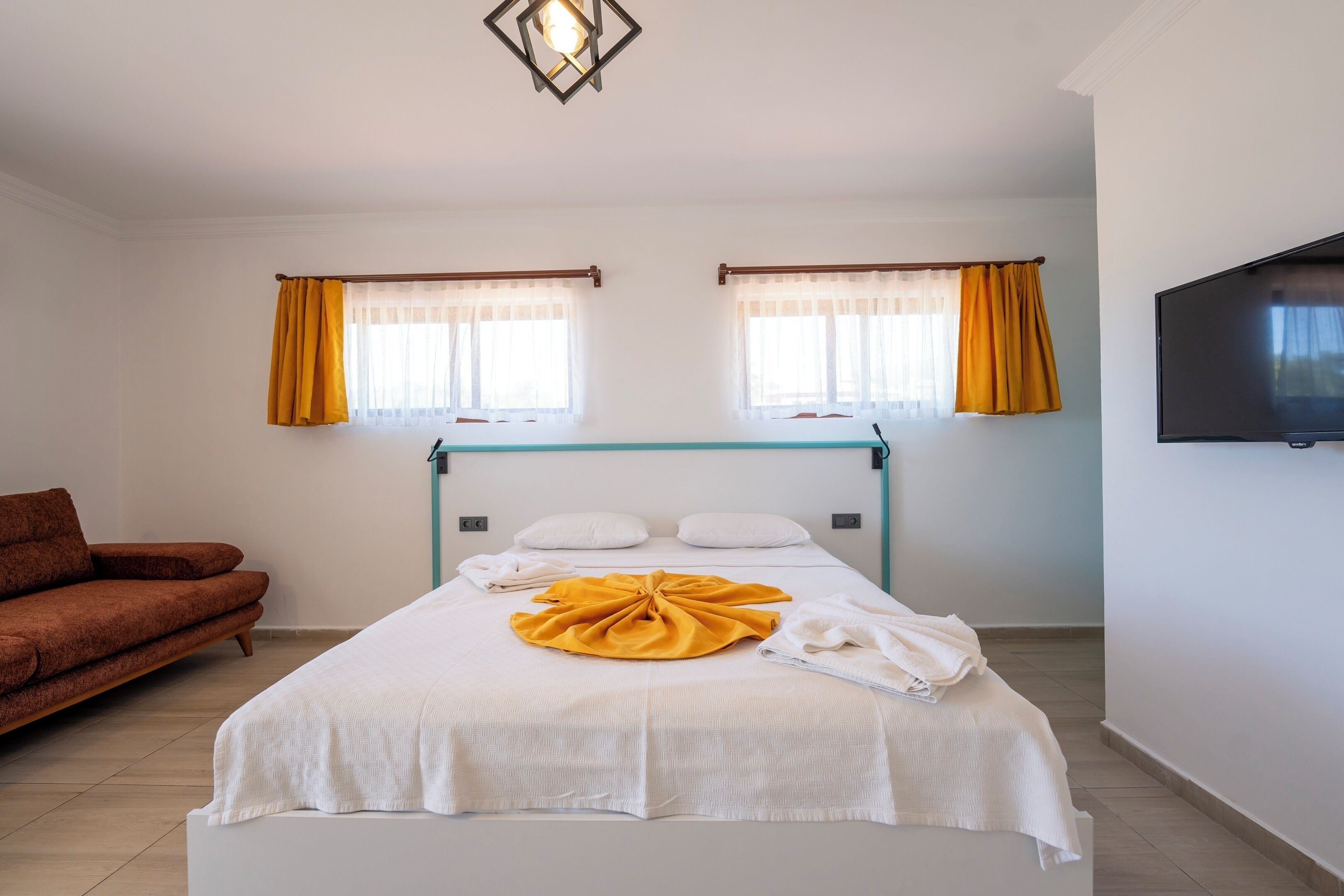 A hotel room with a bed, a sofa, two windows with curtains, a flat-screen TV, and a ceiling light fixture.