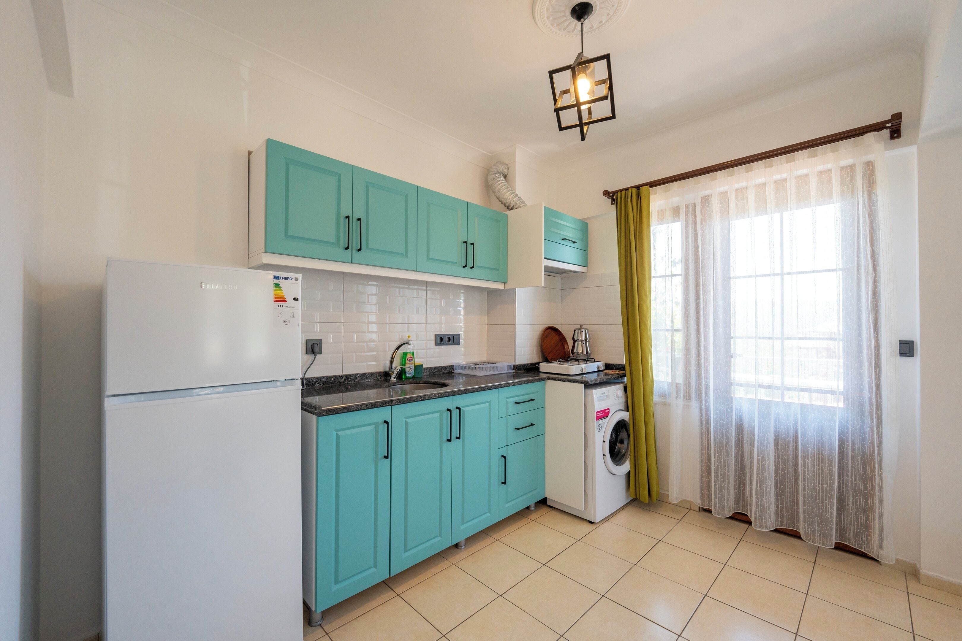 Una cocina con armarios verde mar, un refrigerador blanco, una lavadora y una ventana con cortinas translúcidas.