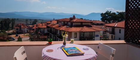Varanda com mesa posta para dois, com vista para área residencial com telhados de terracota e montanhas ao fundo.