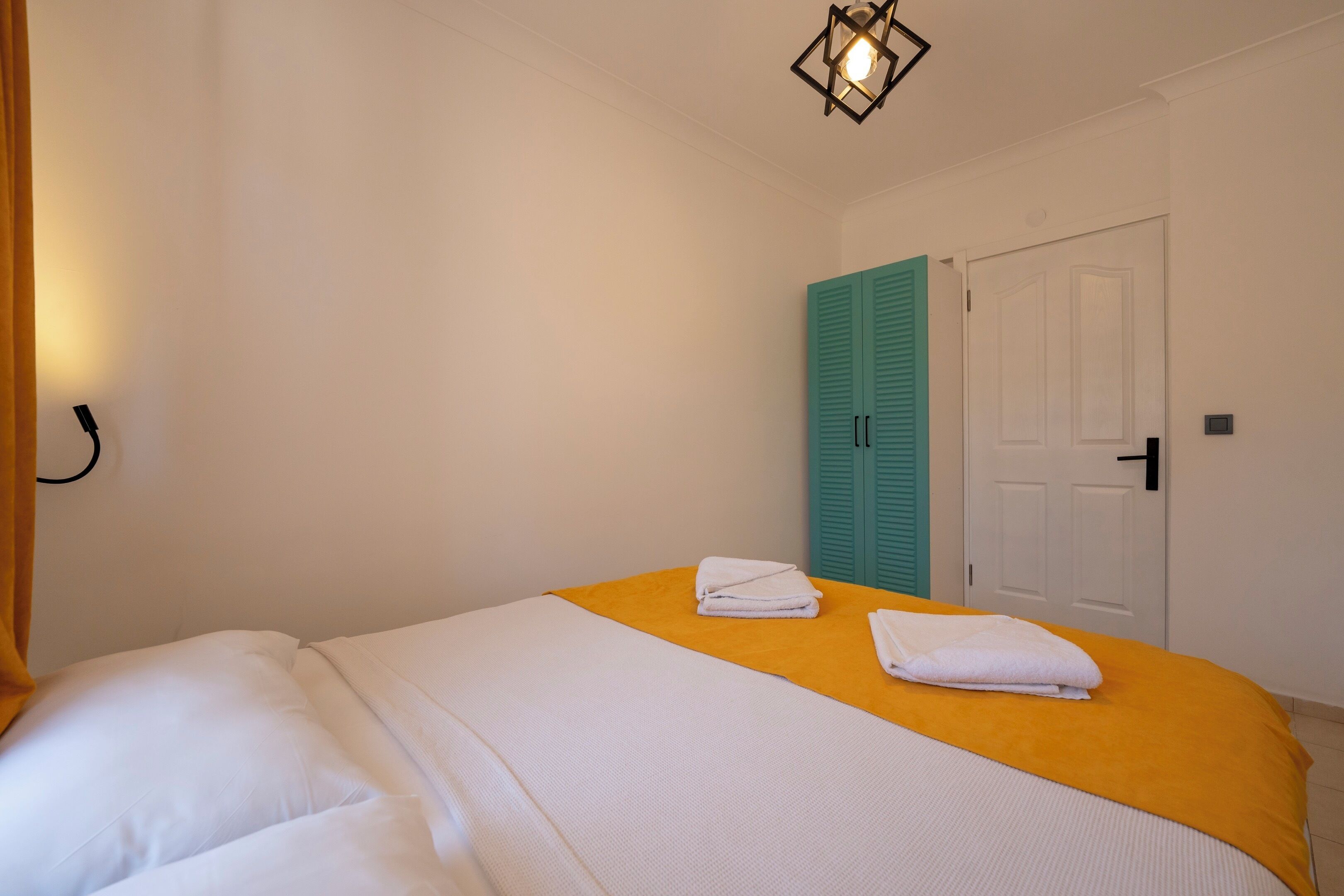 A neatly made bed with white bedding and a yellow accent, a teal door, and a white wall with a light fixture.