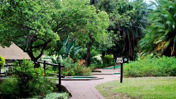 A pathway leading to a mini golf course surrounded by lush greenery and a thatched-roof structure.