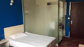 Una habitación con una cama, una mesita, un cubo de basura, una cabina de ducha de vidrio y una puerta.