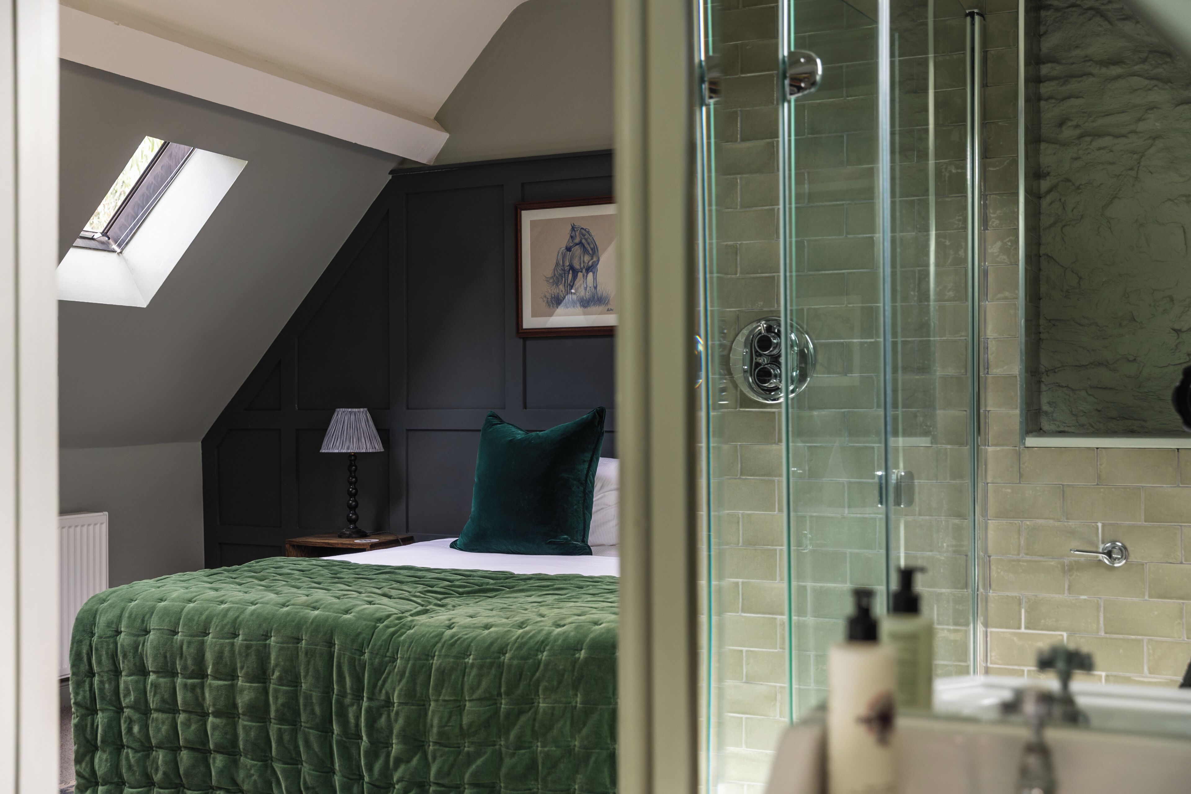 A bedroom with a green bedspread, a nightstand with a lamp, a framed picture on the wall, and a glass shower enclosure.