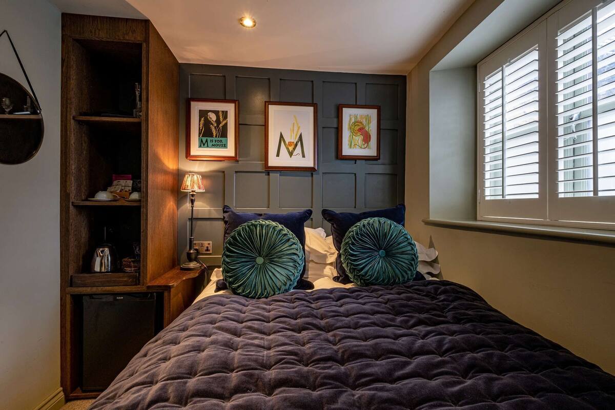 Un dormitorio con cama, mesita de noche, lámpara, estante con libros y cuadros en la pared.