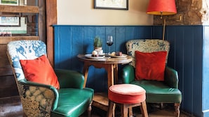 A room with two green upholstered chairs, a small round table with a red cushion, a red floor lamp, and a framed picture on the wall.