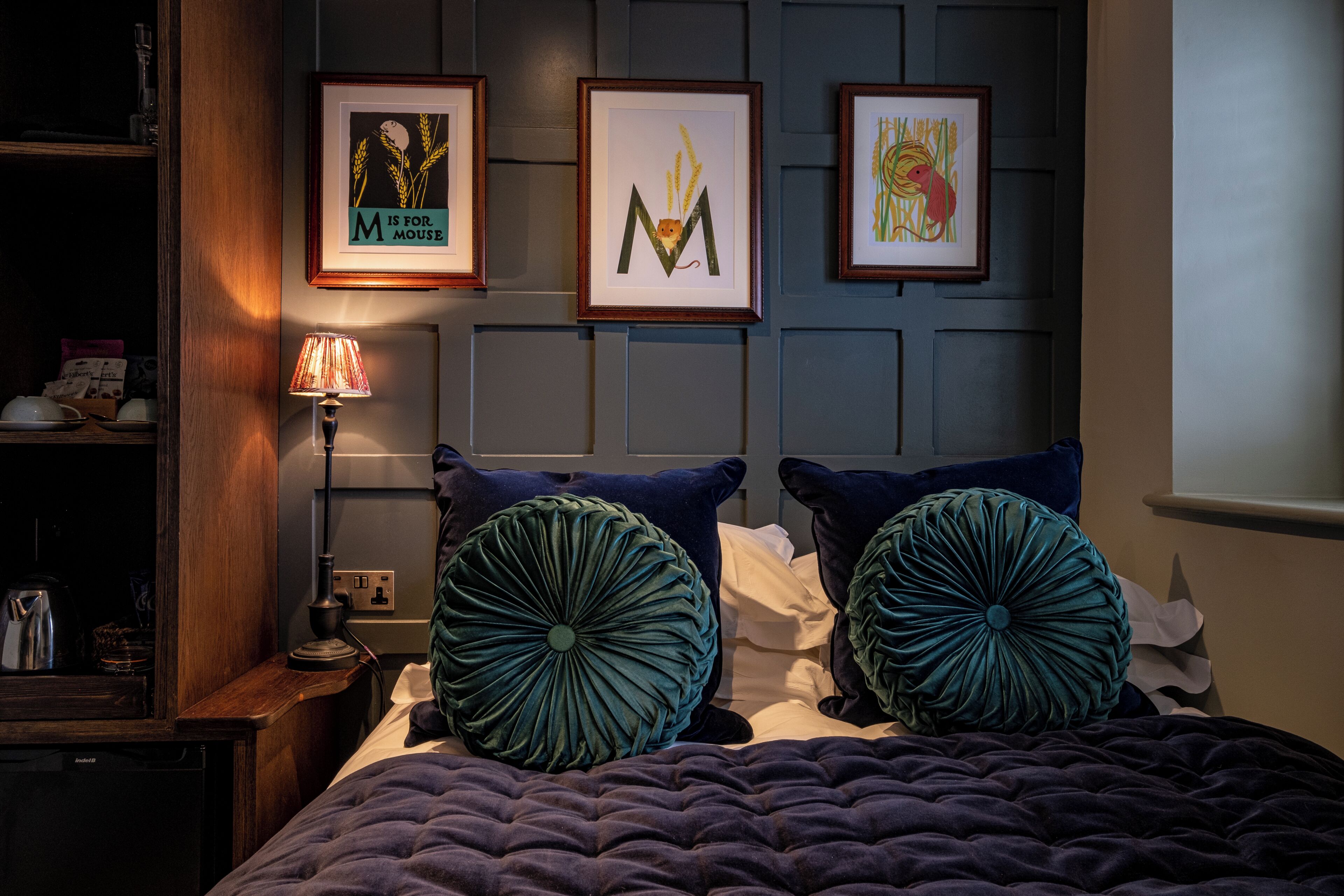 Una cama bien hecha con un edredón azul oscuro, dos almohadas decorativas grandes y una mesita de noche con una lámpara.