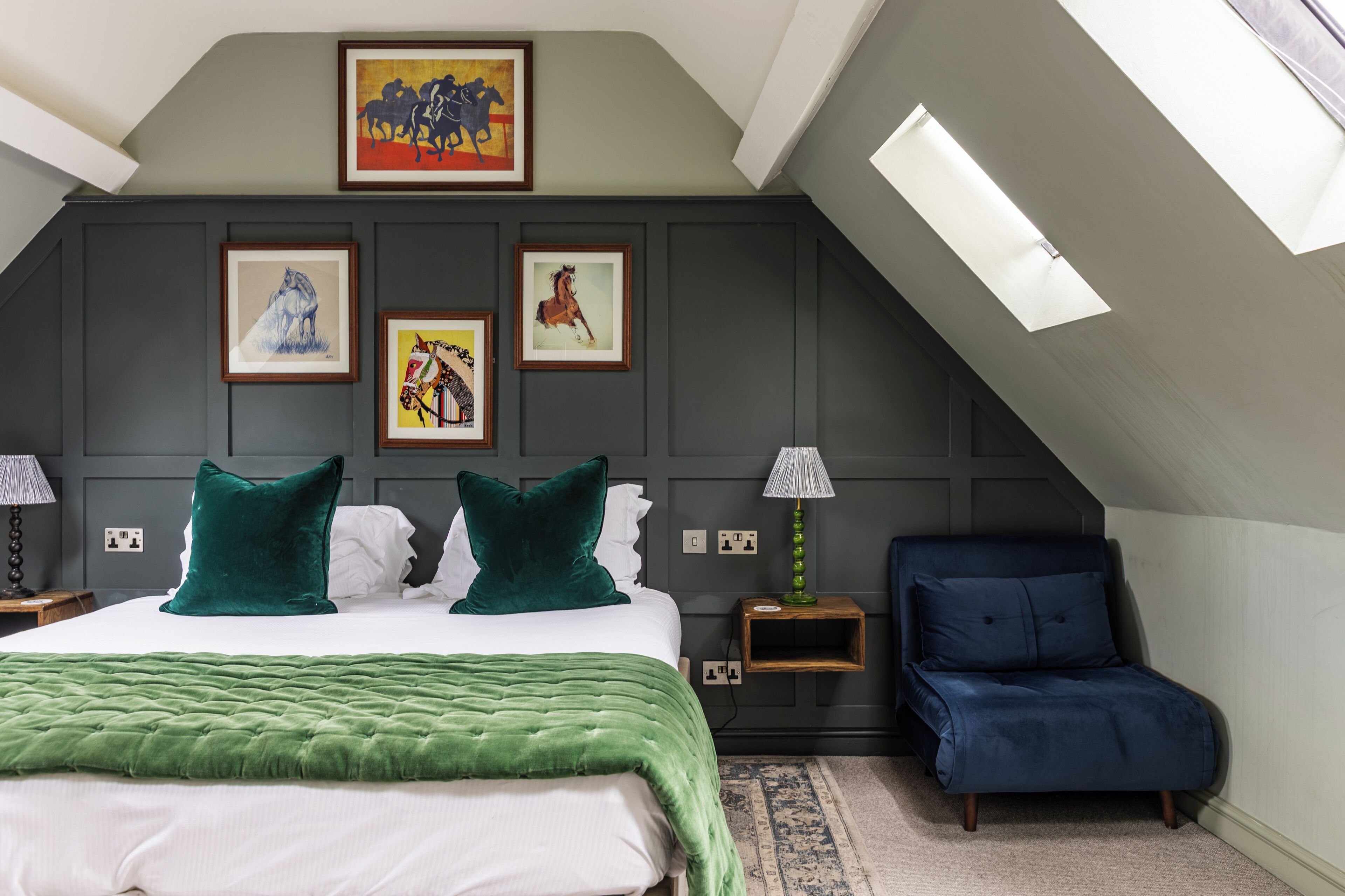 Ein Schlafzimmer mit schrägverdeckter Decke, einem Bett mit grüner Bettwäsche, zwei Kissen, einem blauen Sessel und gerahmten Bildern an der Wand.
