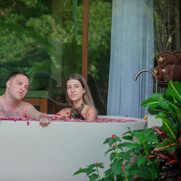 A bathtub filled with rose petals, a towel rack with a towel, and a window with a view of greenery.