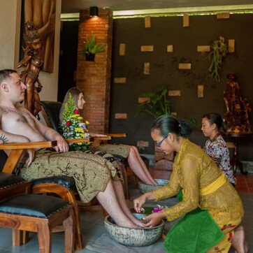 A man seated in a wooden chair with a towel wrapped around his legs, a woman kneeling in front of him, and another woman standing beside them.