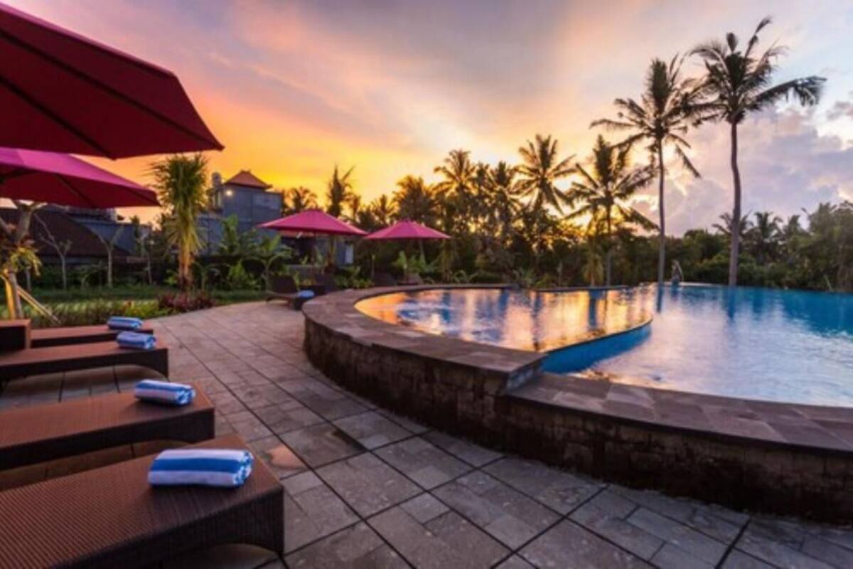A poolside area with lounge chairs and umbrellas, surrounded by palm trees and a sunset sky.
