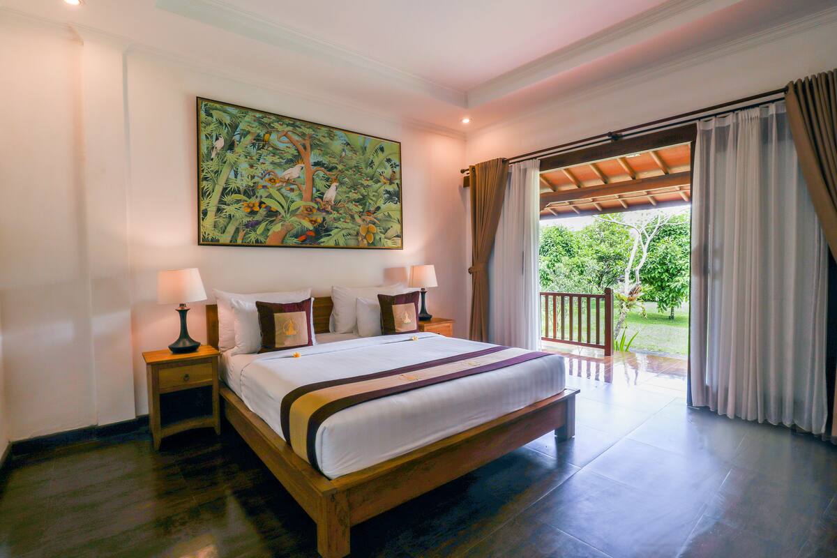 A bedroom with a large bed, two bedside tables, a nightstand, a painting, and a view through a door to an outdoor area.