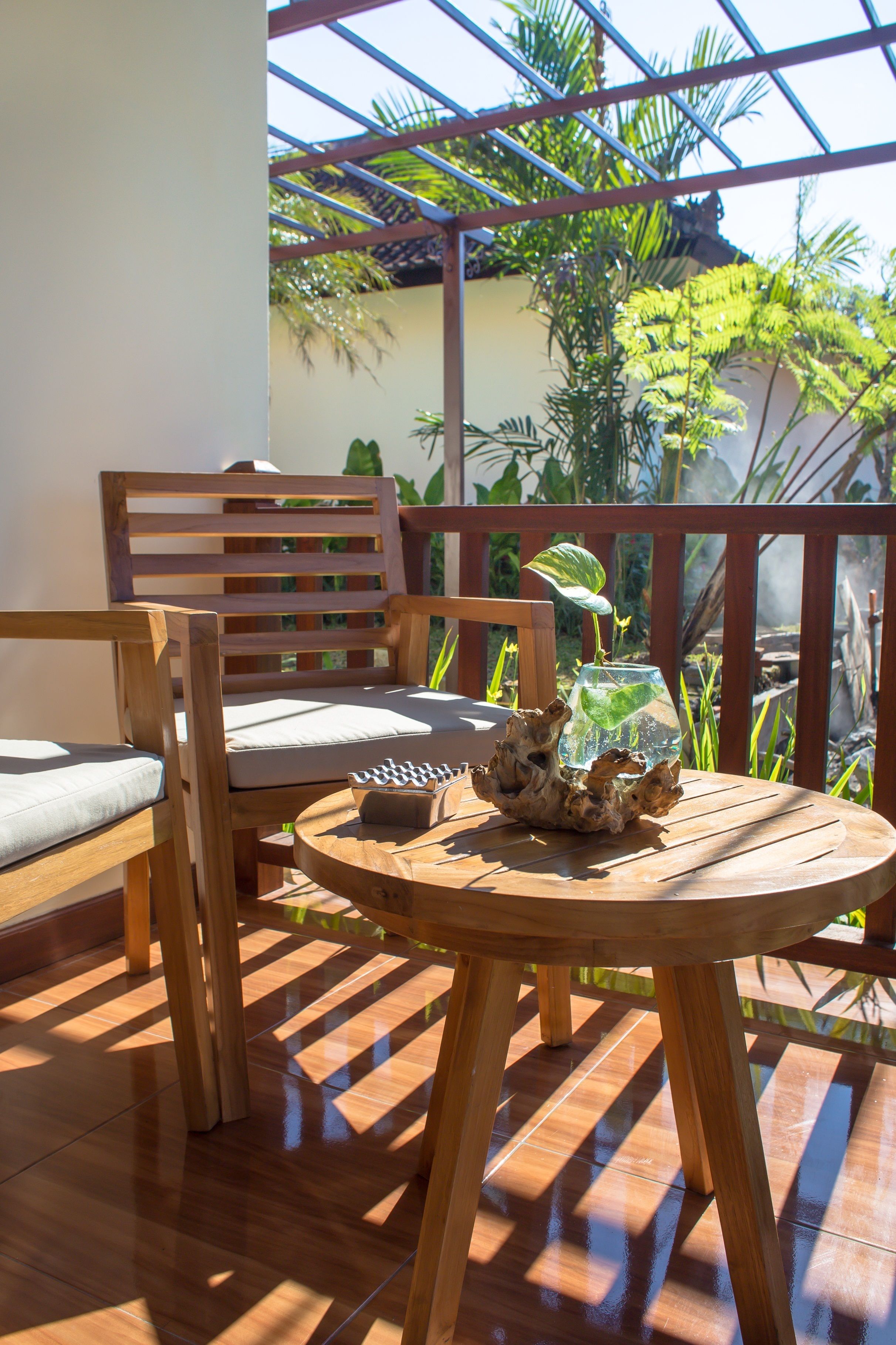 A wooden outdoor seating area with a round table and chairs, a small decorative item on the table, and a view of lush greenery and a building in the background.
