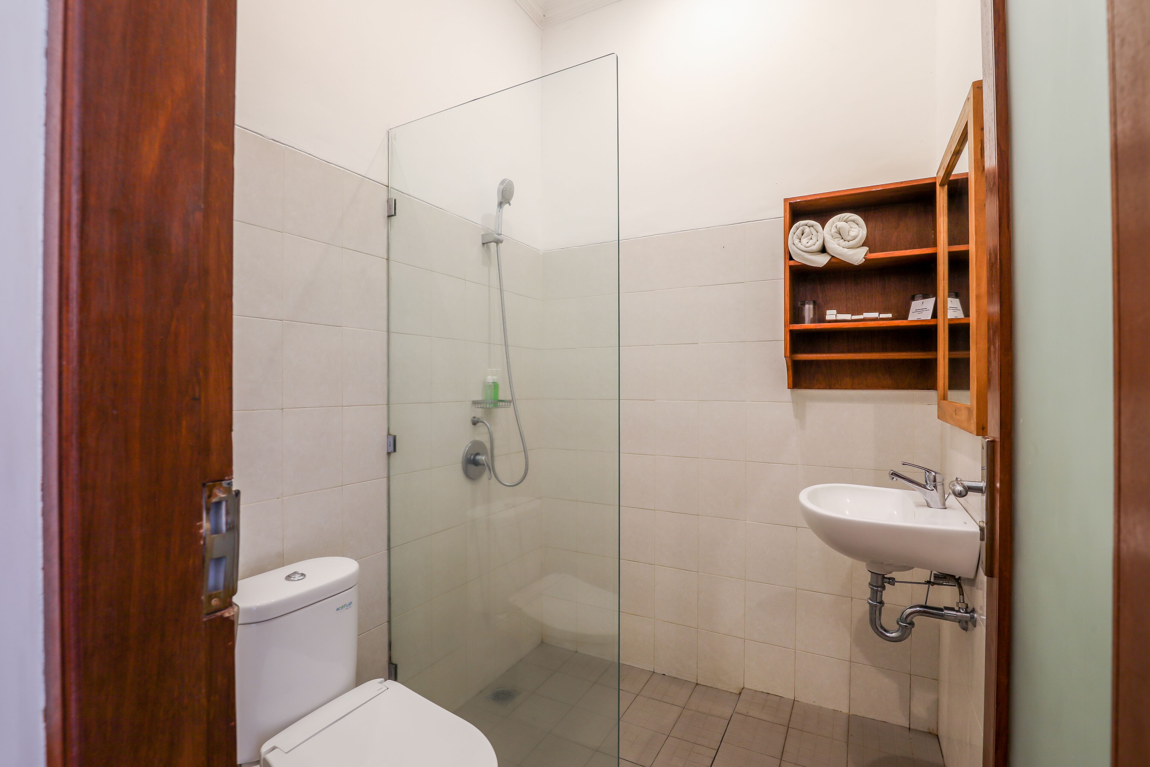A bathroom with a glass-enclosed shower, a toilet, and a sink with a wooden shelf above it.