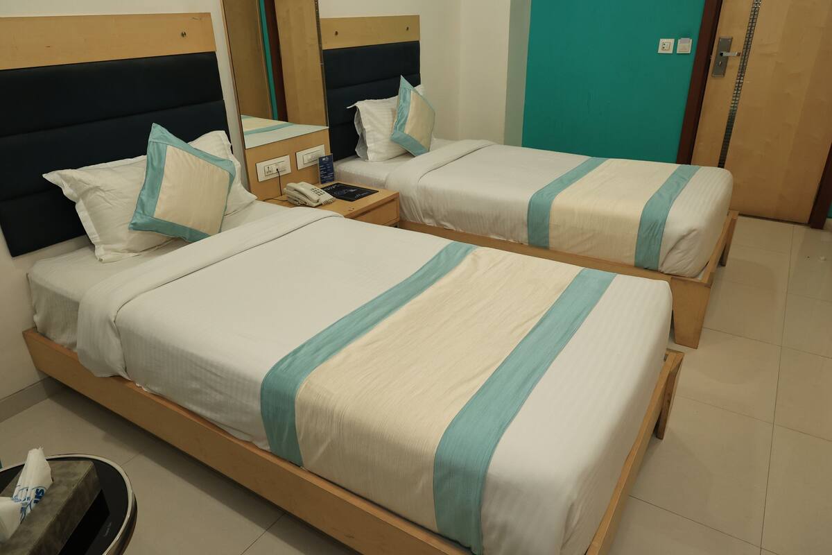 A hotel room with two single beds, each with a wooden frame and white bedding with teal stripes. There is a bedside table with a phone and a small lamp. The room has a wooden headboard and a teal accent wall.