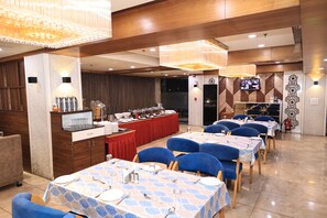 A restaurant with tables set for dining, a buffet area, and a television screen. - Hotel SN Blu (Ahmedabad)