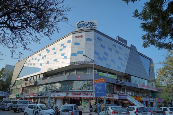 A modern hotel with a distinctive geometric facade, multiple ground-level shops, and a clear blue sky. - Hotel SN Blu (Ahmedabad)