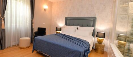 Une chambre d’hôtel avec un grand lit, deux tables de chevet avec des lampes, un pouf rond et une fenêtre avec des rideaux.
