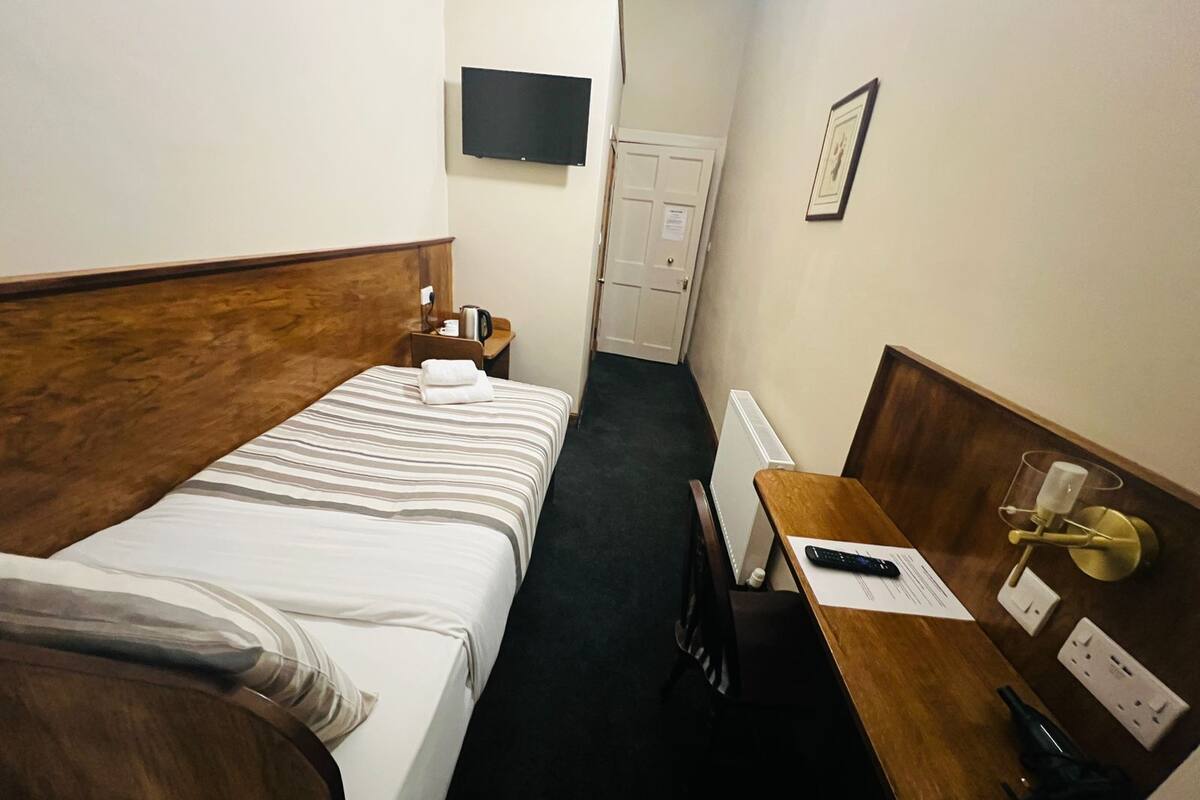Ein kleines Hotelzimmer mit einem Einzelbett, einem Holz-Kopfteil, einem Nachttisch mit Lampe, einem Schreibtisch mit Stuhl, einem an der Wand montierten Fernseher und einer Tür zu einem Badezimmer.