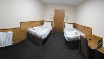 Habitación de hotel estrecha con dos camas individuales, un radiador, una mesita con una tetera y tazas, y un televisor montado en la pared.