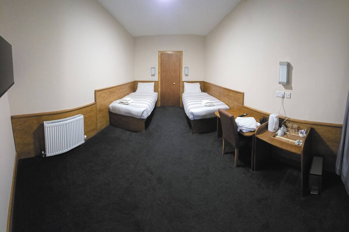 Ein Hotelzimmer mit zwei Einzelbetten, einem Schreibtisch mit Stuhl, einem Heizkörper, einem an der Wand montierten Fernseher und einem Fenster mit Vorhängen.