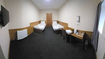 Une chambre d’hôtel avec deux lits simples, un bureau avec une chaise, un radiateur, un téléviseur fixé au mur et une fenêtre avec des rideaux.