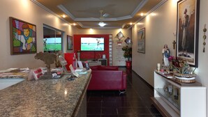 Zona de recepción con mostrador, asientos rojos, un televisor mostrando un evento deportivo y varias decoraciones en la pared, incluyendo pinturas y objetos religiosos.