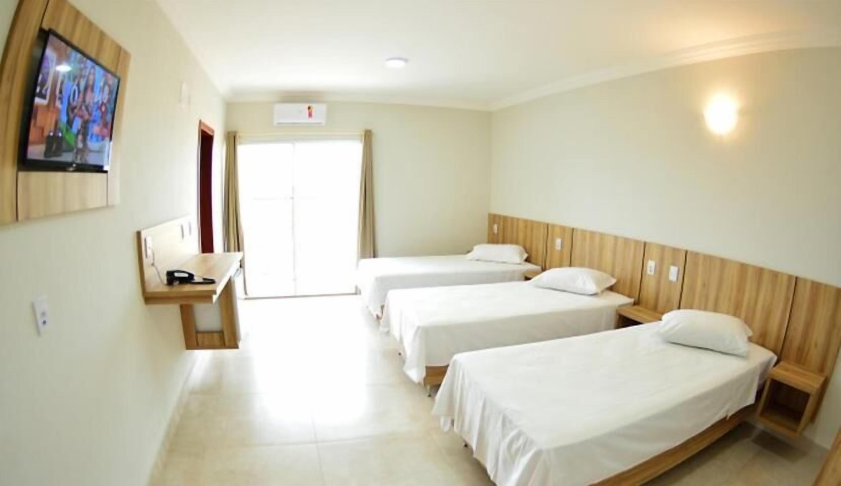 A hotel room with three single beds, a wooden headboard, a desk with a chair, and a television mounted on the wall.