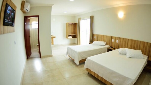 A hotel room with two beds, a wooden headboard, a bedside table with a lamp, and a TV mounted on the wall. - Dallas Palace Hotel (Rio Paranaiba)
