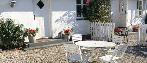 A white building with a black roof, a white door, and windows. There is a white fence, a gravel area with a white round table and four white chairs.