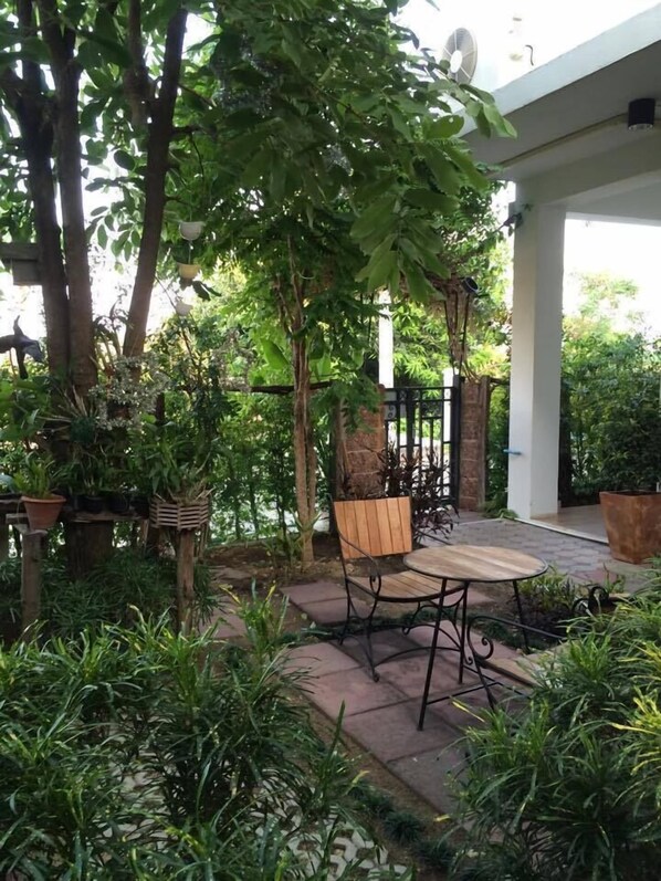 Un área de patio al aire libre con una mesa y sillas de madera, rodeada de abundante follaje y plantas en macetas.