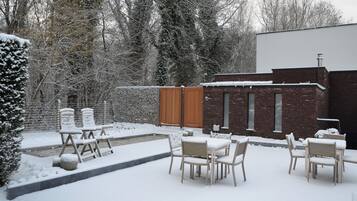 Een besneeuwde terras buiten met een bakstenen muur, houten omheining en meerdere stoelen en tafels.