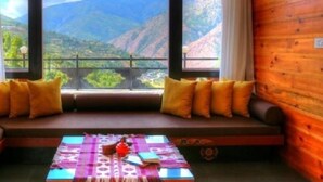 A wooden cabin with a large window overlooking a mountainous landscape. Inside, there is a cozy seating area with cushions and a table with books and a teapot. - Dewa Thimphu by Realm (Thimphu)