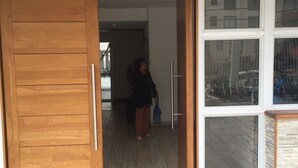 Eine Holztür führt in einen Raum mit Fliesenboden, einem Fenster mit Blick auf Fahrräder und einer Fußmatte mit einem Logo.