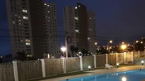 Ein Schwimmbecken mit einer gefliesten Terrasse und Metallleitern. Weiße Liegestühle und ein Zaun mit Tor sind vorhanden. Im Hintergrund sind hohe Gebäude zu sehen.