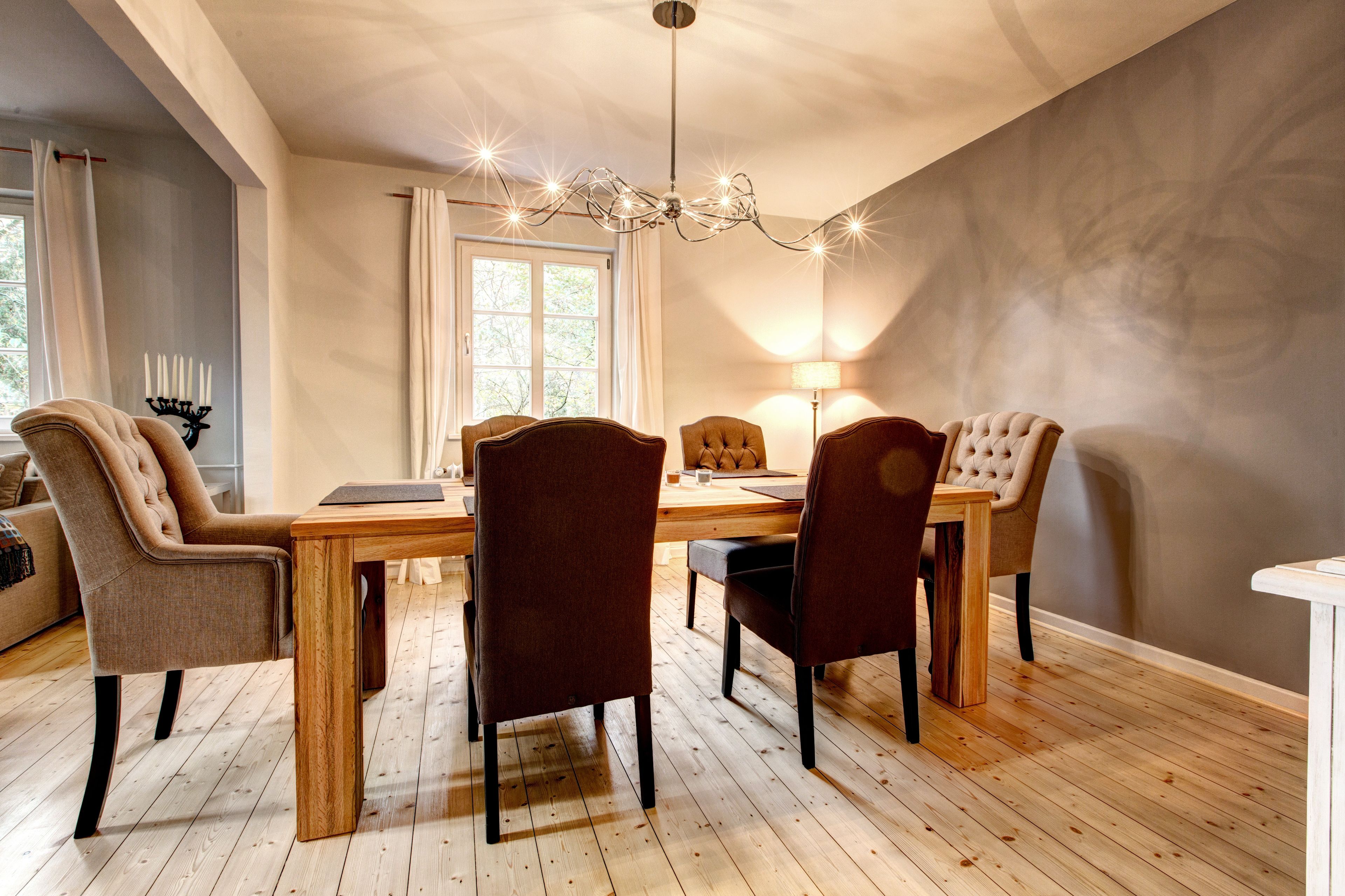 A dining area with a wooden table and four chairs, a chandelier, and a window with curtains.