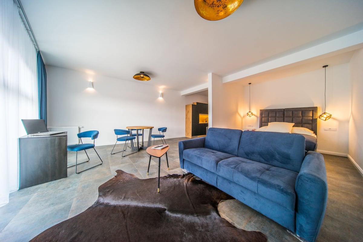 A modern living room with a blue sofa, a wooden coffee table, and a leather rug. There is a dining area with blue chairs and a small table.