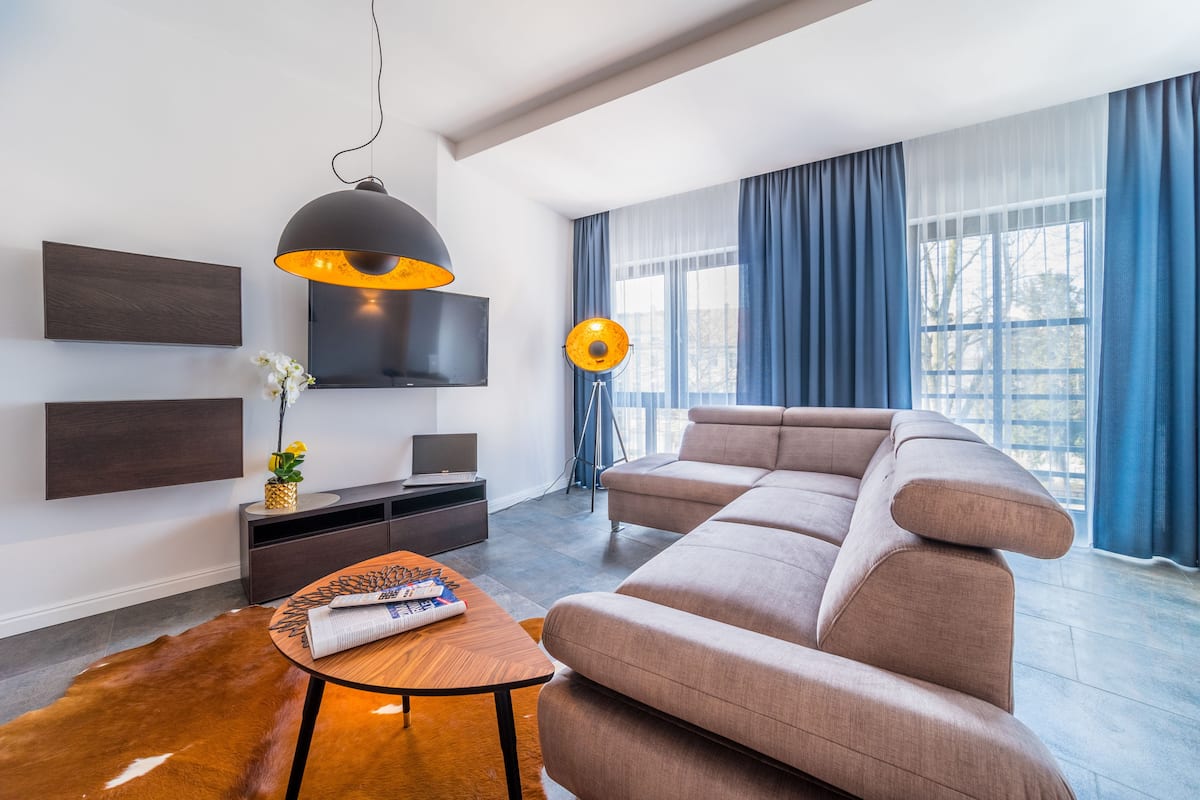 A modern living room with a sectional sofa, a round coffee table, a TV mounted on the wall, and a large window with blue curtains.