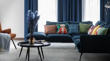 Een moderne woonkamer met een donkerblauwe bank, meerdere kleurrijke kussens, een klein rond tafeltje met een vaas en een hoge vloerlamp.