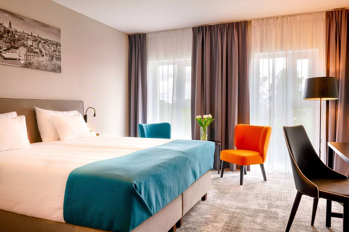 Une chambre d’hôtel avec un grand lit, un bureau, deux chaises et une fenêtre avec des rideaux.