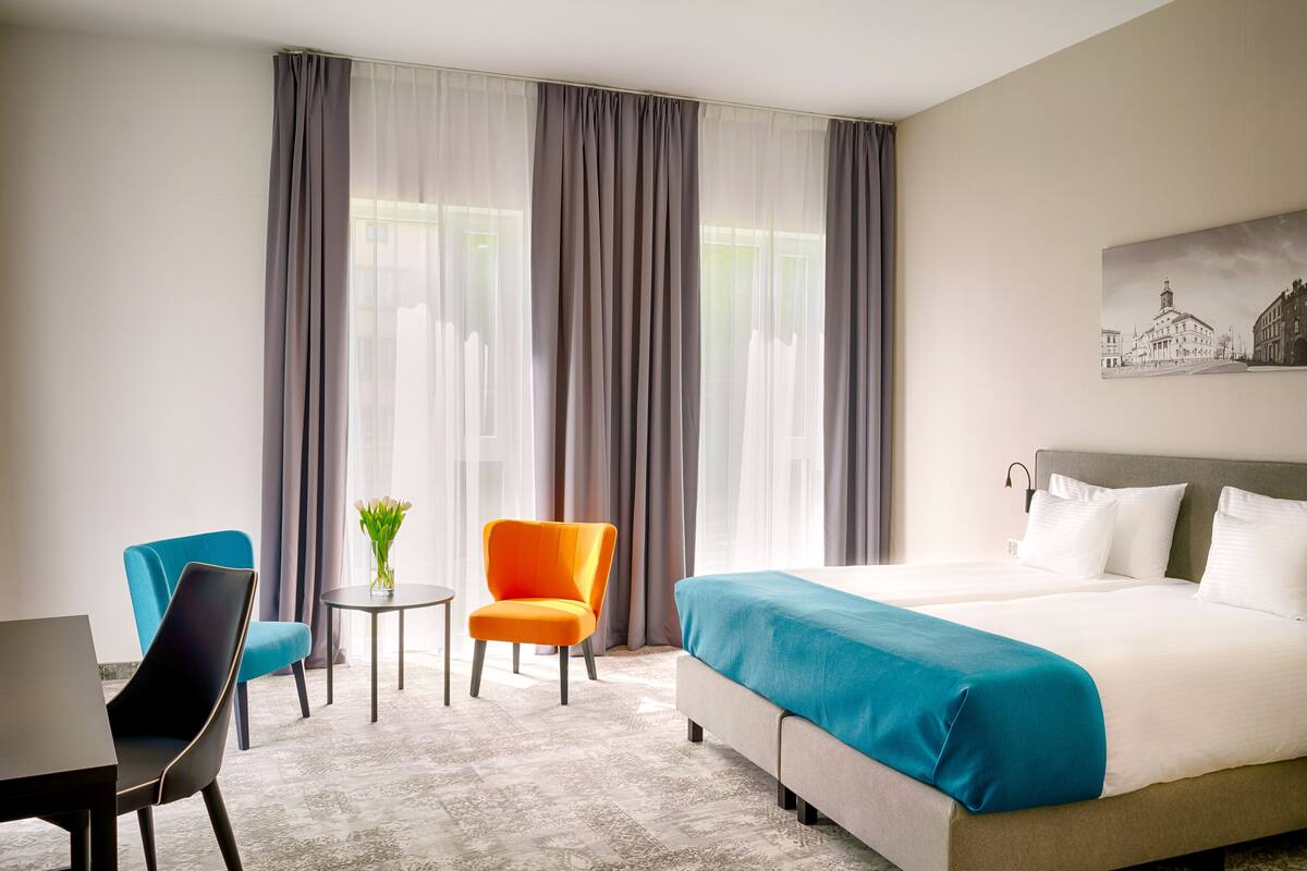 Une chambre d’hôtel avec un grand lit, deux chaises, une petite table et une fenêtre avec des rideaux.