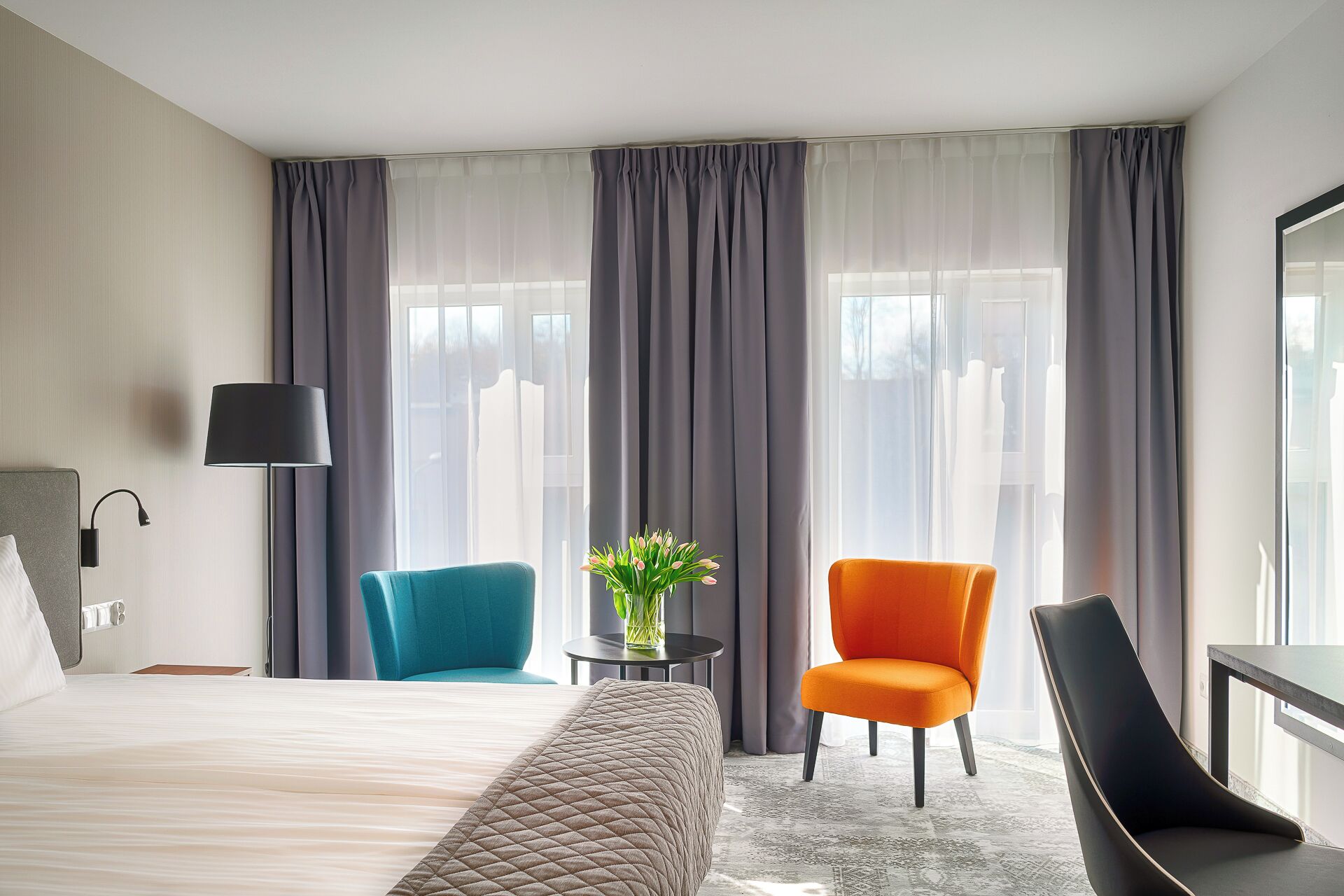 A hotel room with a bed, two chairs, a table with a vase, and a lamp.