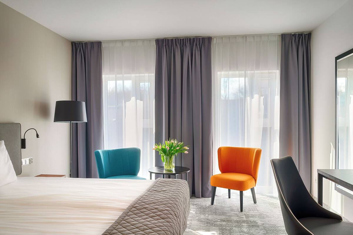 Une chambre d’hôtel avec un lit, deux chaises, une table avec un vase et une lampe.