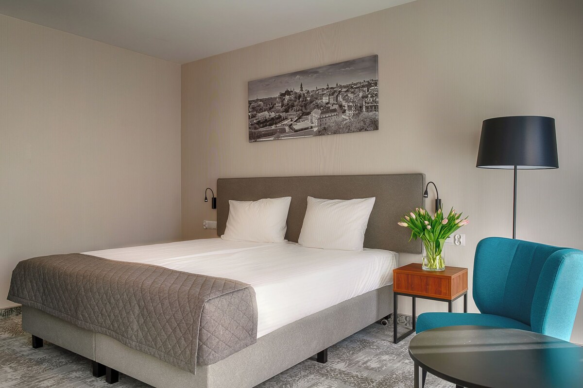 A hotel room with a bed, a nightstand, a lamp, a chair, and a cityscape painting on the wall.
